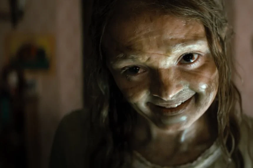 Close-up de uma jovem (Katie) com um sorriso largo, perturbador e não natural, olhando diretamente para a câmera em uma cena dimly lit de A Maldição da Múmia (2026). Sua pele parece cerosa e úmida.