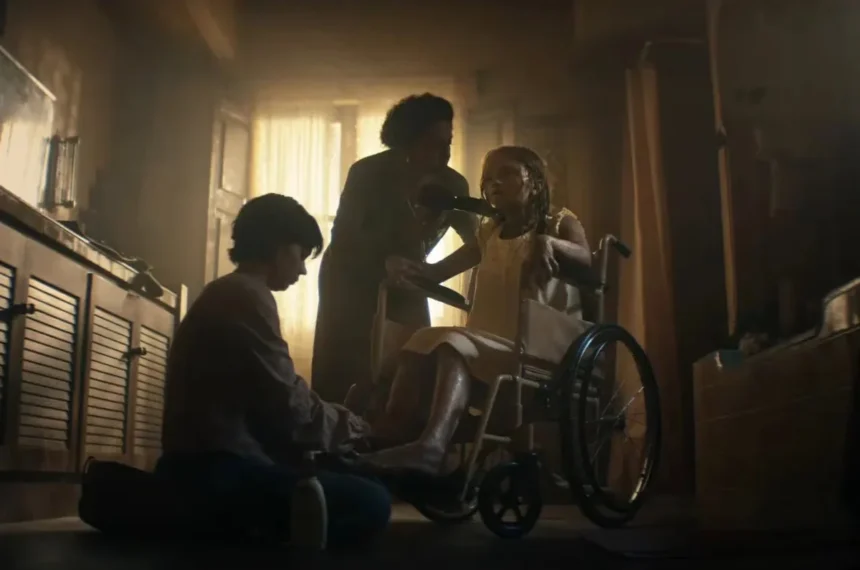 Três pessoas em um quarto escuro e empoeirado: uma menina pálida sentada em uma cadeira de rodas sendo cuidada por um adulto ajoelhado e outra mulher ao fundo em A Maldição da Múmia (2026).