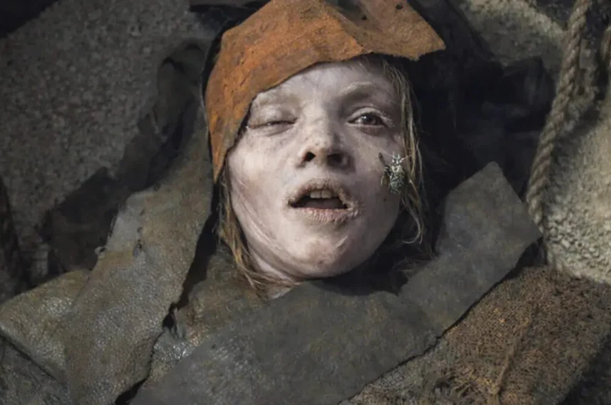 Rosto pálido e acinzentado de uma menina deitada entre faixas de tecido antigo, com um besouro rastejando em sua bochecha no filme A Maldição da Múmia (2026).