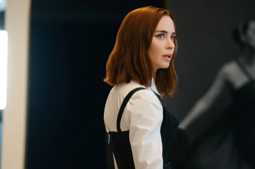 Emily Blunt como Emily Charlton em cena de "O Diabo Veste Prada 2", vestindo camisa branca e corset preto com olhar atento sobre o ombro.