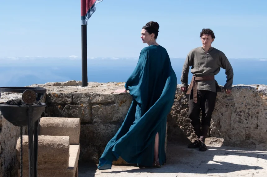 Anne Hathaway vestida com uma túnica azul ao lado de Tom Holland em um terraço de pedra rústico com vista para o mar azul no filme A Odisseia.