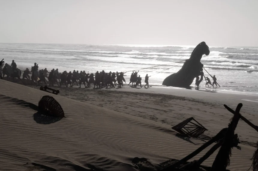 Imagem em contraluz mostrando a silhueta de dezenas de soldados na areia da praia puxando com cordas um colossal Cavalo de Troia de madeira pelas águas rasas.