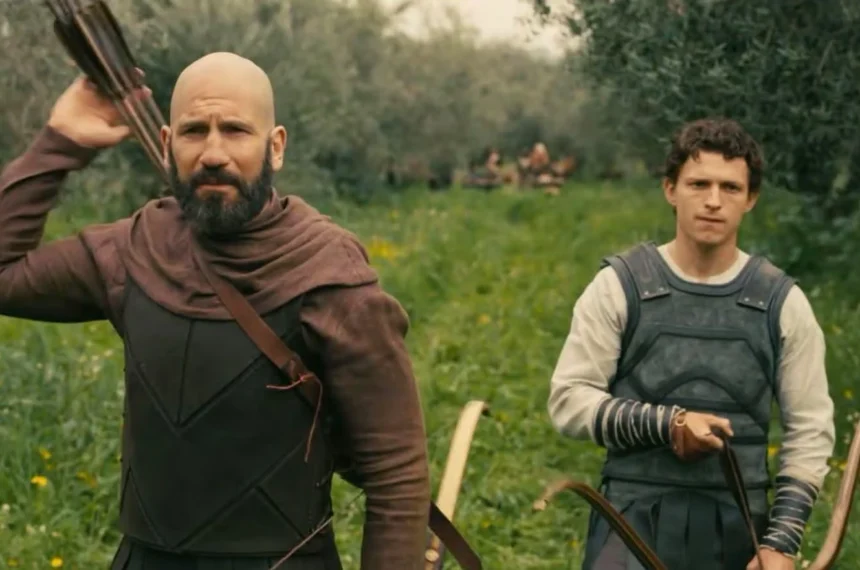 Jon Bernthal e Tom Holland caminham por um campo verde segurando arcos de caça em cena do filme A Odisseia, de Christopher Nolan.