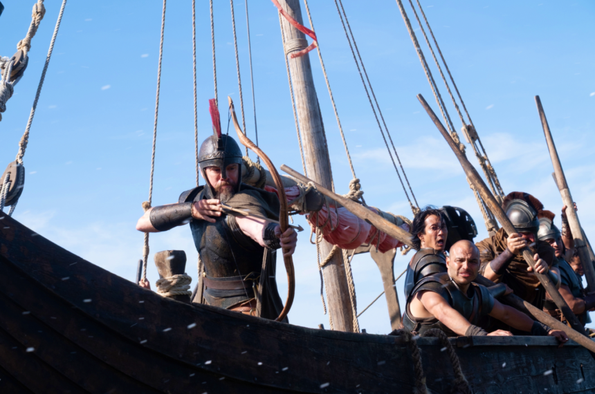 Matt Damon de armadura e capacete puxando a corda de um arco em um barco de madeira balançando no mar, com outros marinheiros remando freneticamente ao fundo.