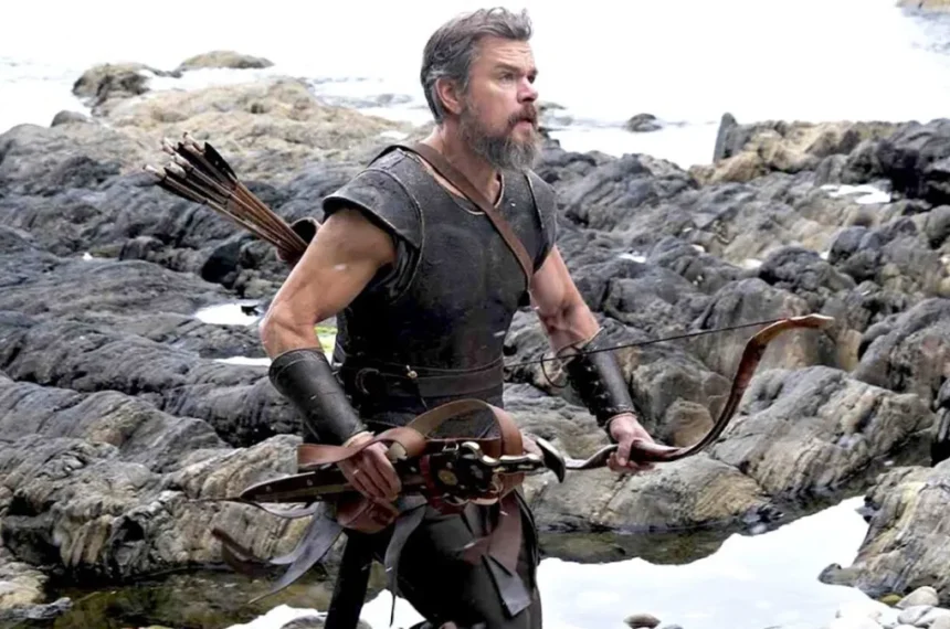Matt Damon vestido com armadura escura de couro, segurando um arco de madeira e flechas nas costas, caminhando por rochas escuras na beira do mar.
