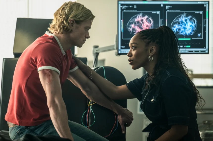 Naomi Ackie conforta Tom Rhys Harries, que está sentado num laboratório médico com uma ferida severa no braço. Monitores com exames cerebrais estão visíveis ao fundo.