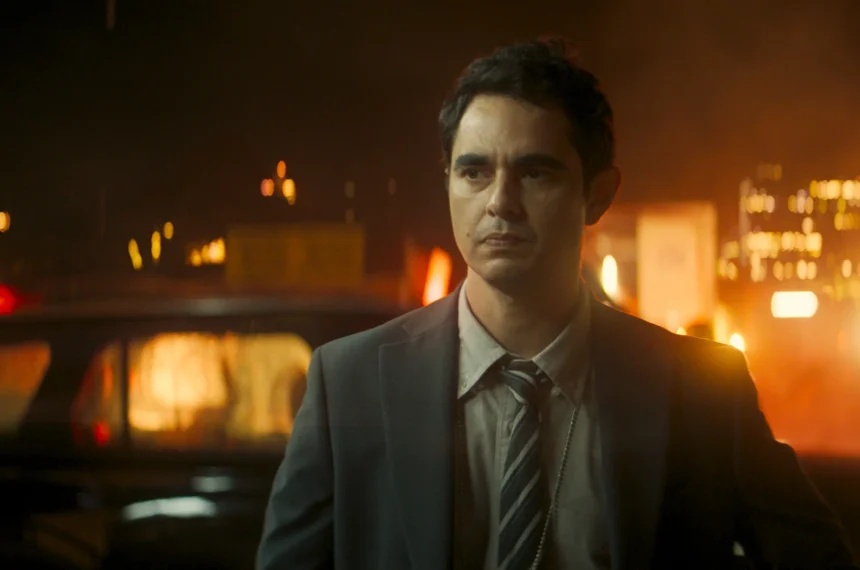 O ator Max Minghella, interpretando o Detetive John, caminha com uma expressão séria por um cenário noturno iluminado por chamas e luzes de veículos da polícia.