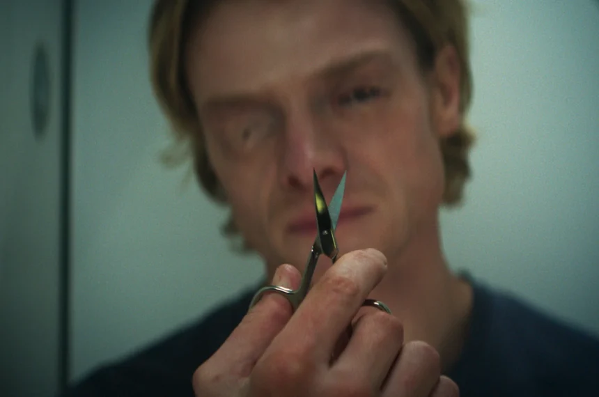 Tom Rhys Harries, com um olho esbranquiçado e o rosto deformado, segura uma pequena tesoura entre os dedos, com uma expressão perturbada.